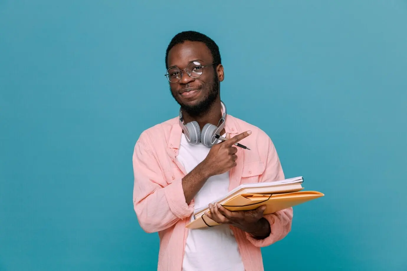 Selbstbewusster junger Afroamerikaner, der Bücher und einen Stift hält, mit Kopfhörern um den Hals, isoliert vor blauem Hintergrund.